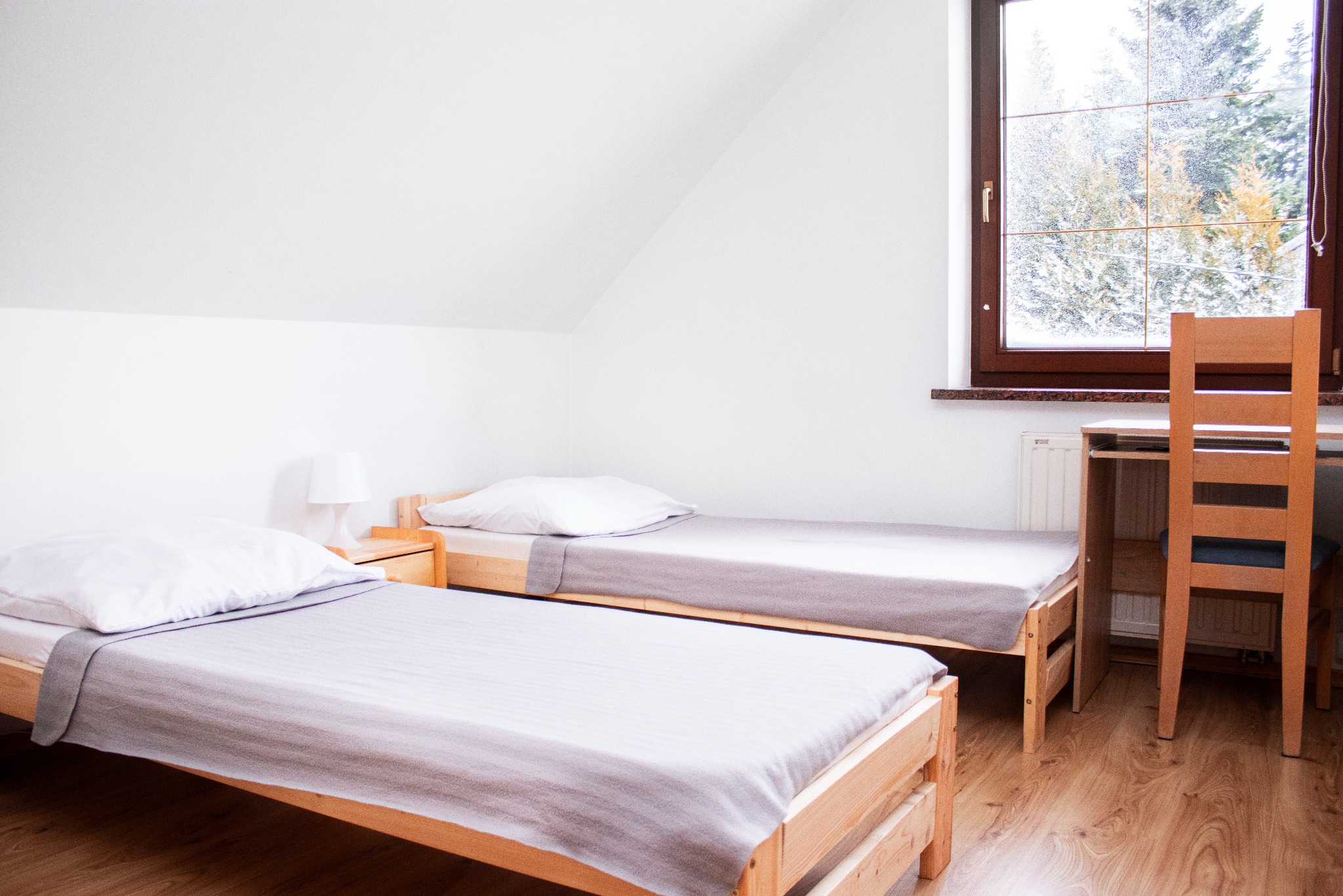 Pokój z dwoma pojedynczymi łóżkami z drewnianymi ramami i szarą pościelą, biurko z krzesłem pod oknem z widokiem na zaśnieżone drzewa.