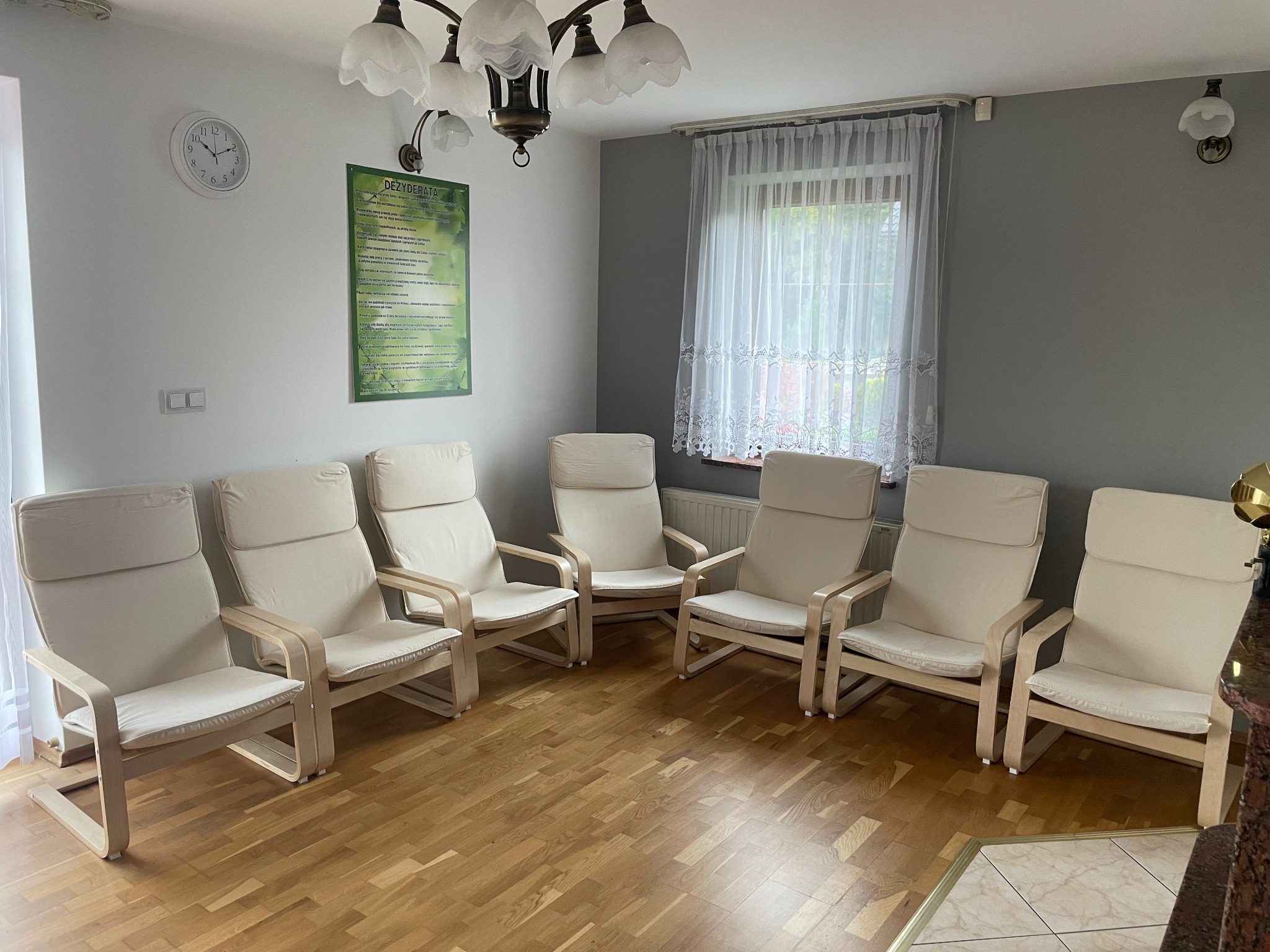 Jasne, drewniane krzesła z beżowymi poduszkami ustawione w okręgu na parkiecie w jasnym pomieszczeniu terapeutycznym, z widocznym zegarem i plakatem na ścianie.
