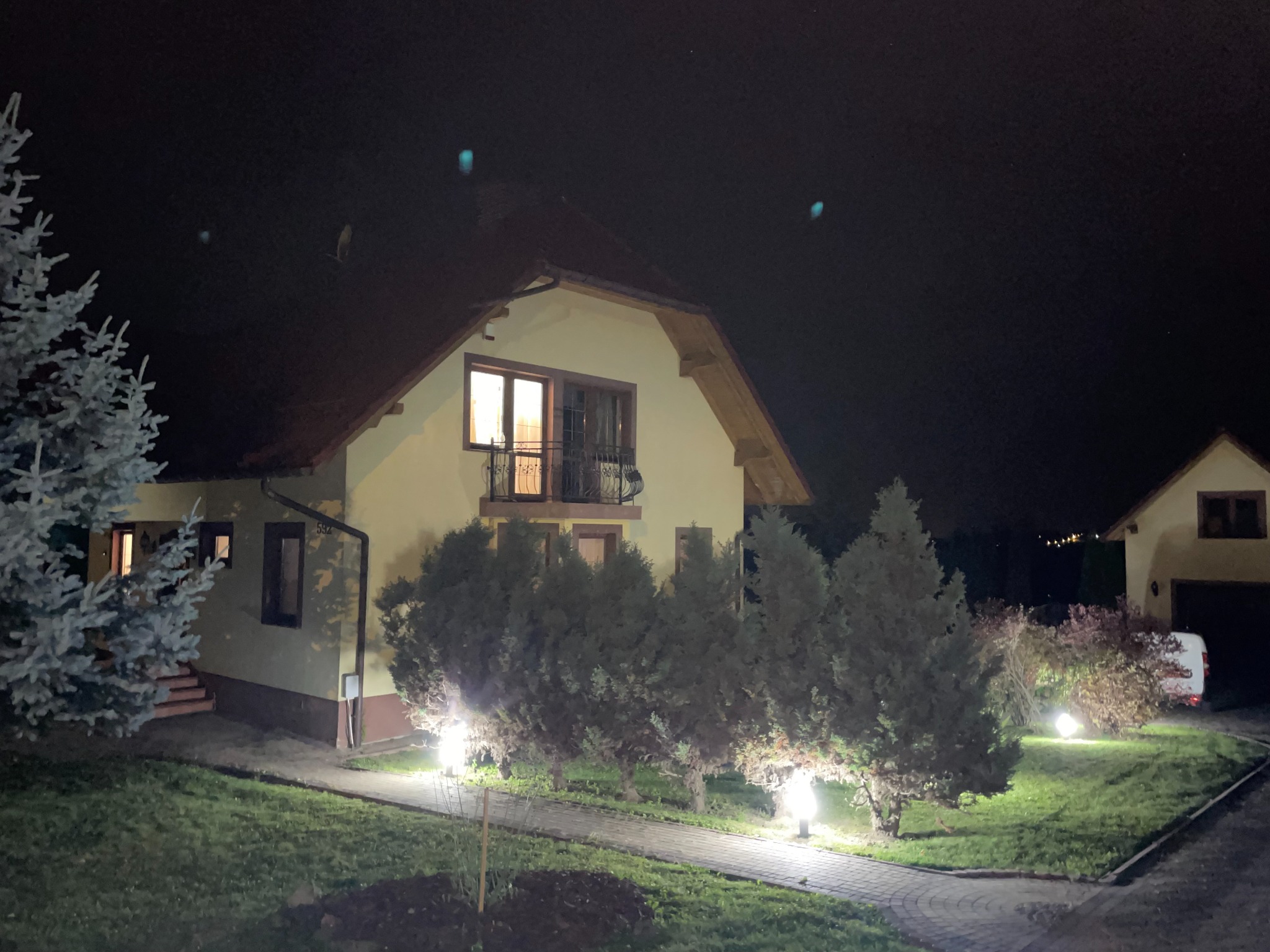 Nocna fotografia domu jednorodzinnego z oświetlonym ogrodem i ścieżką z kostki brukowej, widok z poziomu trawnika.