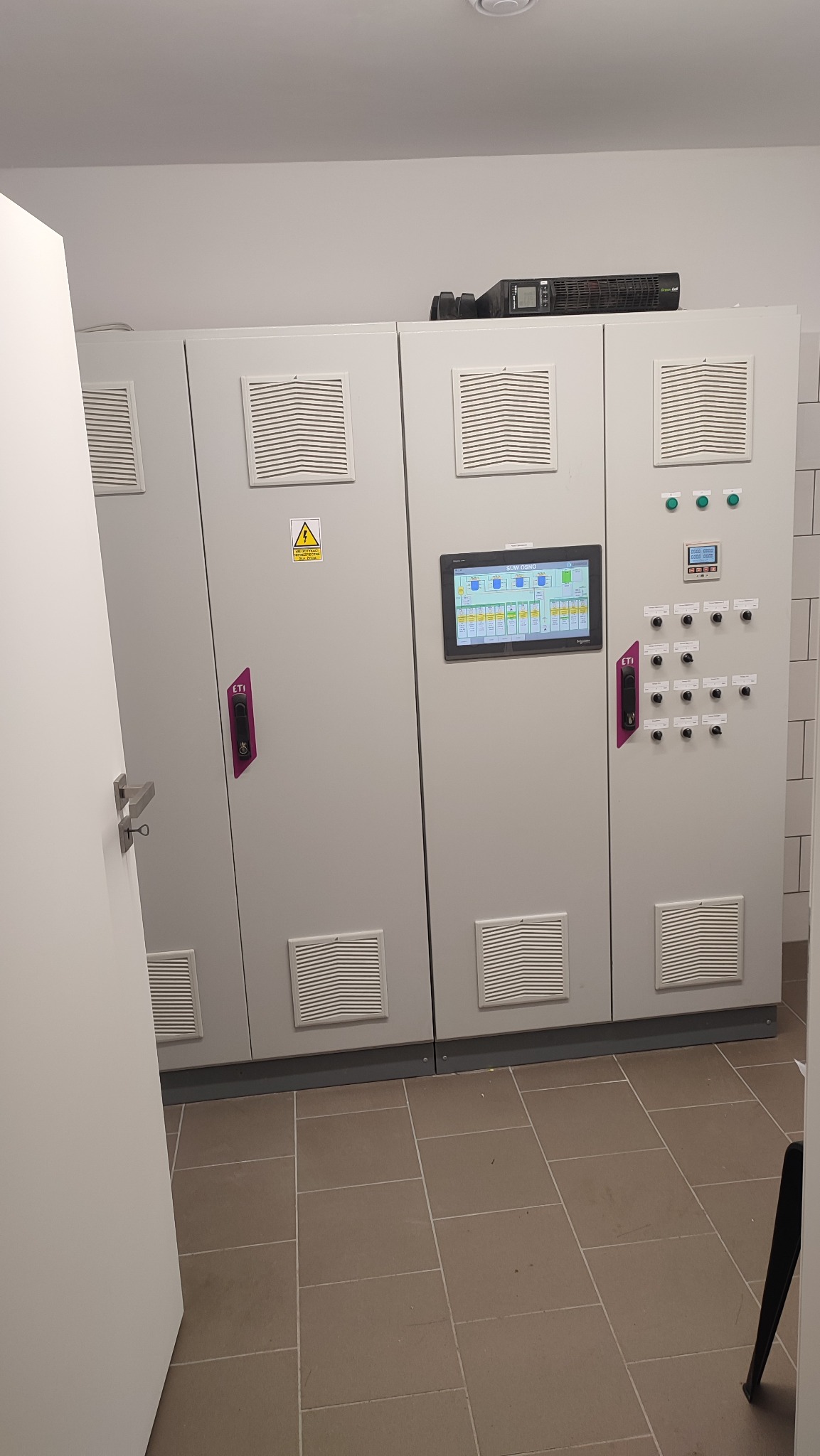 Szafa sterownicza z panelem dotykowym i elementami sterującymi, widoczny UPS na górze, w pomieszczeniu technicznym.