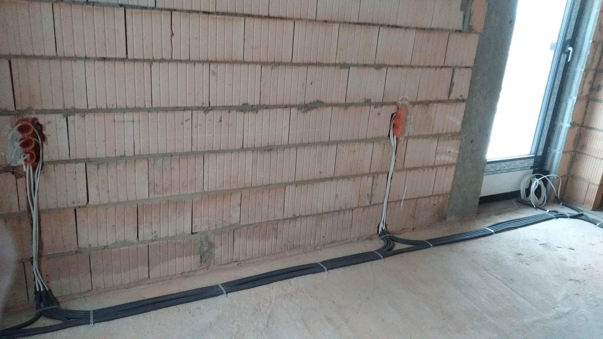 Wewnętrzna instalacja elektryczna w surowym stanie: przewody w szarych peszlach ułożone wzdłuż ściany z pustaków ceramicznych, wychodzące z puszek instalacyjnych z trzema otworami, widoczne okno...