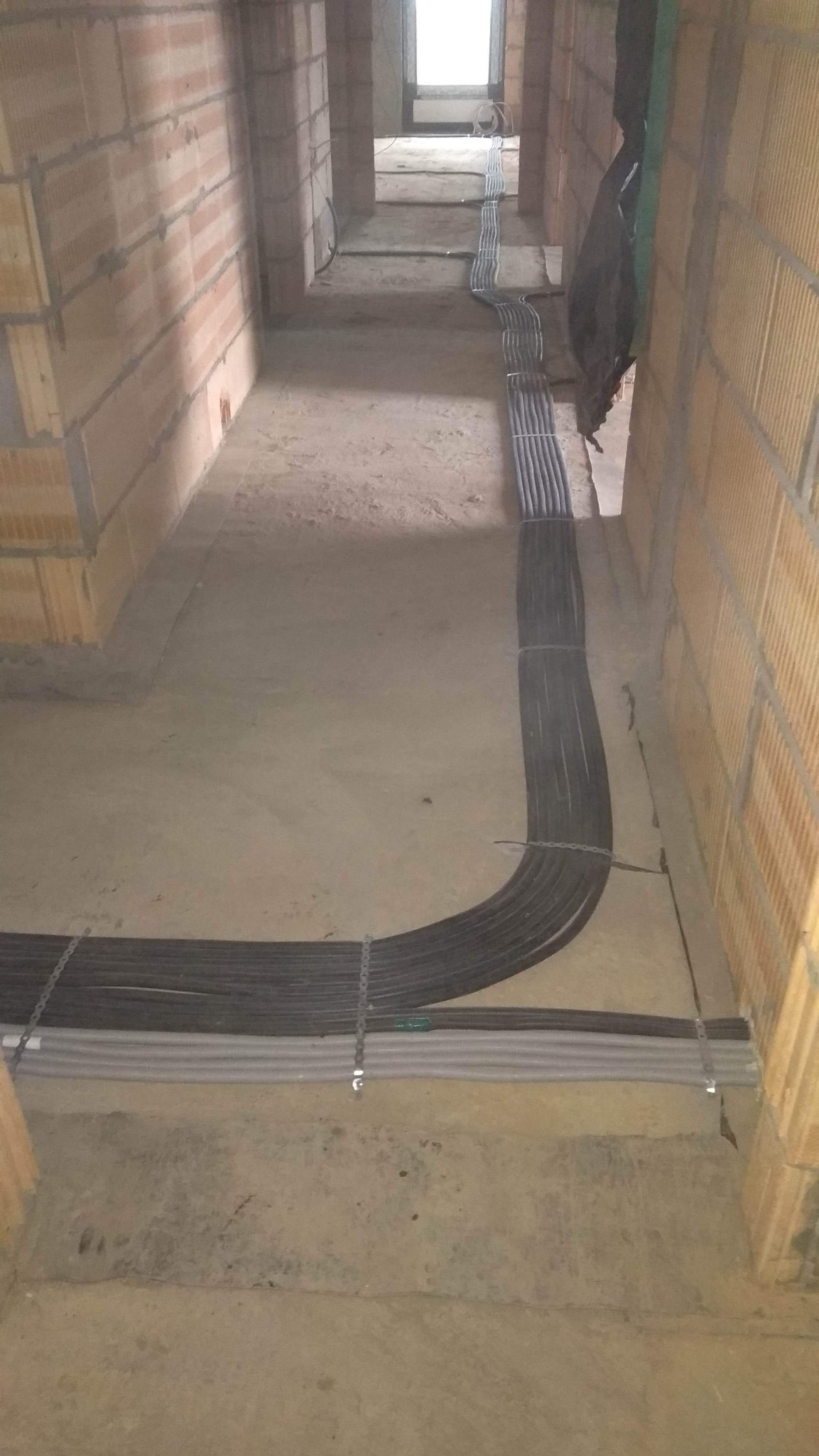 Wiązka kabli elektrycznych prowadzona w korytarzu budynku w trakcie budowy, zabezpieczona metalowymi opaskami, widoczne ceglane ściany i surowy beton