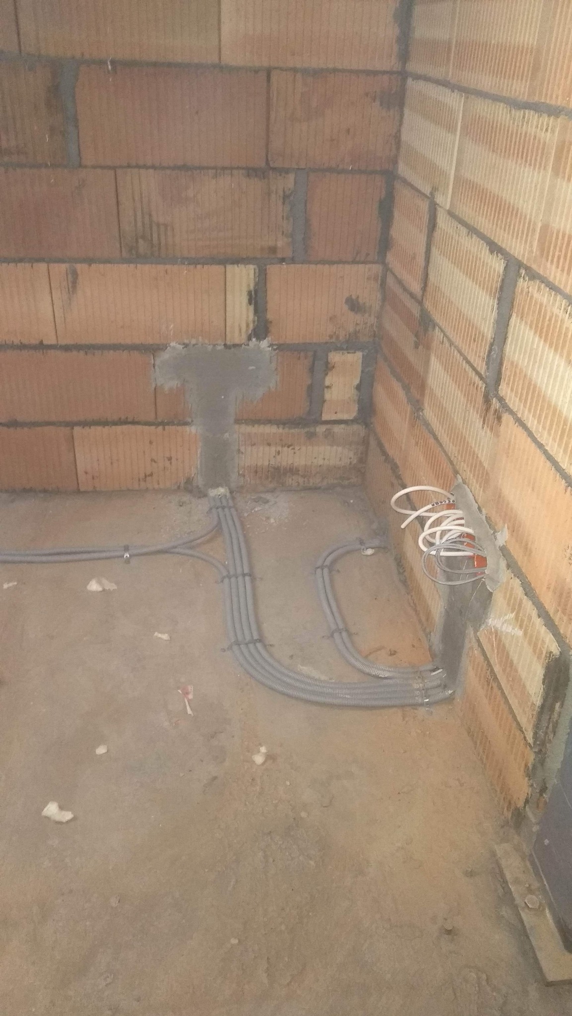 Wewnętrzny róg pomieszczenia z surowymi ścianami z czerwonej cegły, w którym widoczne są szare, elastyczne rury instalacji elektrycznej prowadzone po podłodze i wchodzące w ścianę, gdzie wystają...