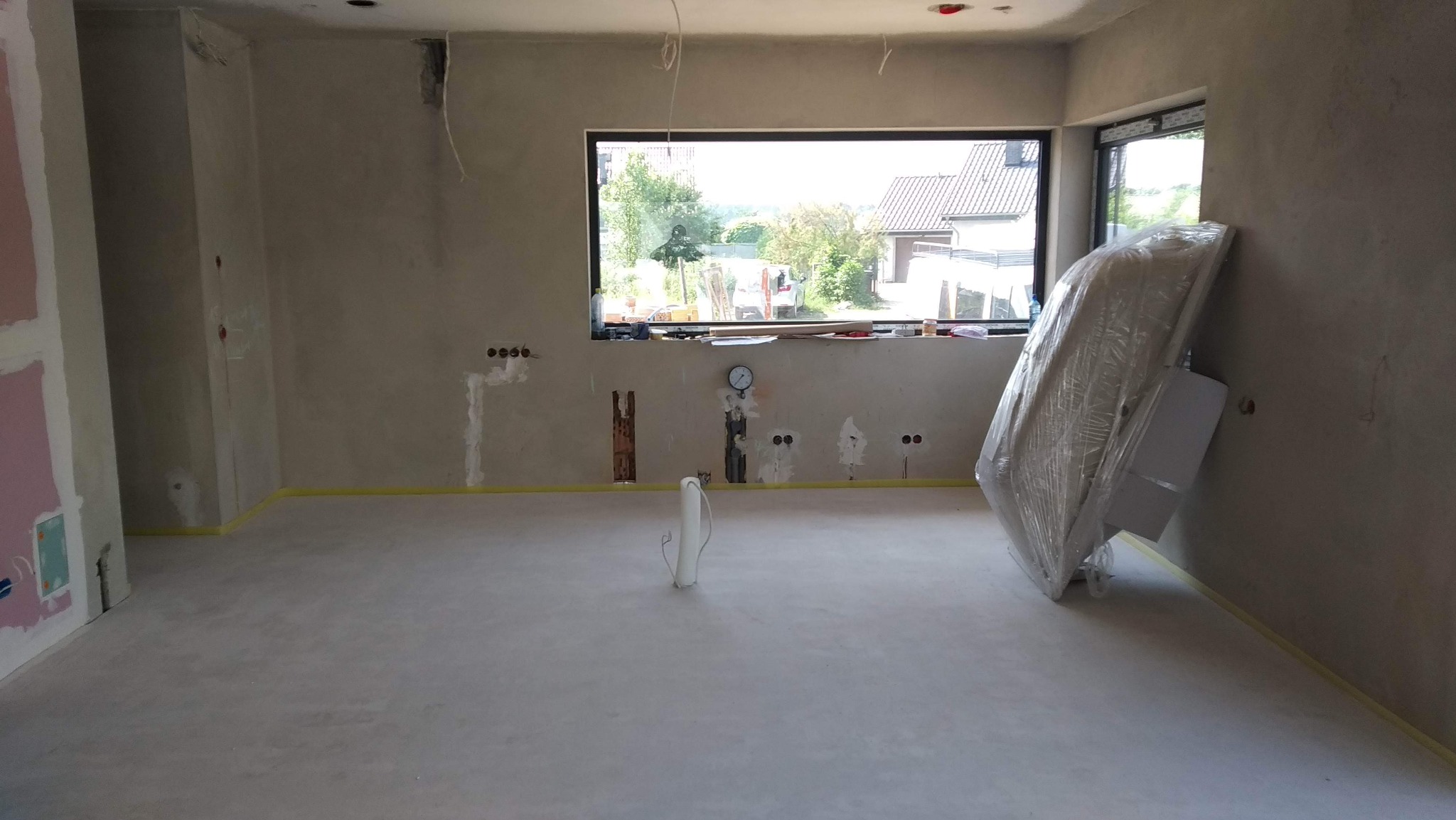 Surowe wnętrze w trakcie remontu: widoczne przygotowane podłoże pod posadzkę, okablowanie elektryczne w ścianach, żółta taśma zabezpieczająca, okno z widokiem na domy i zieleń, oraz sprzęt AGD...