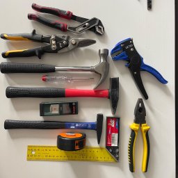 Jakub Lada - Zestaw narzędzi warsztatowych na białym stole: kombinerki, młotki, miarka zwijana, kątownik, szczypce do zdejmowania izolacji, poziomica laserowa Bosch Zamo, nożyce do blachy i wkrętak.