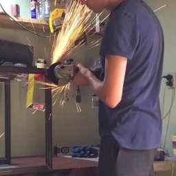 Jakub Lada - Pracownik w okularach ochronnych i słuchawkach tnący metalową konstrukcję szlifierką kątową, iskry w warsztacie.