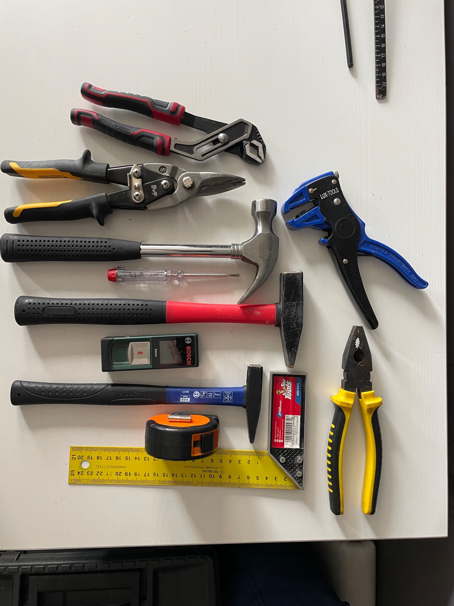 Zestaw narzędzi warsztatowych na białym stole: kombinerki, młotki, miarka zwijana, kątownik, szczypce do zdejmowania izolacji, poziomica laserowa Bosch Zamo, nożyce do blachy i wkrętak.