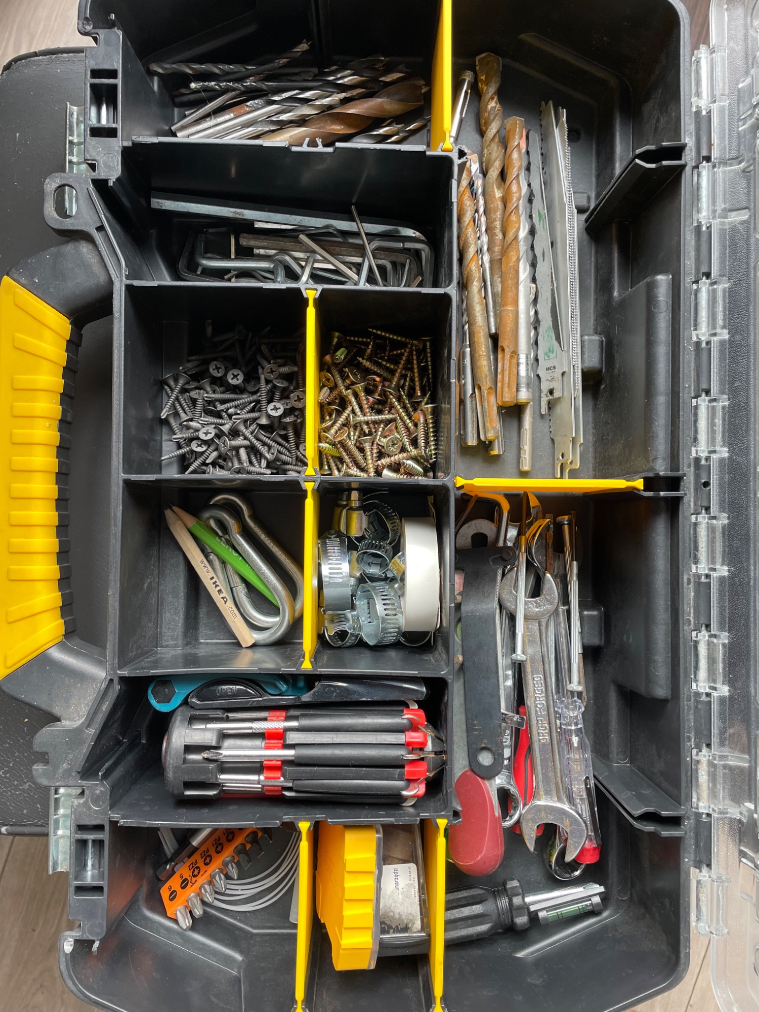 Otwarta skrzynka narzędziowa z przegródkami, zawierająca wiertła, śruby, klucze, wkrętaki i inne akcesoria, w tym karabińczyki i ołówek IKEA.