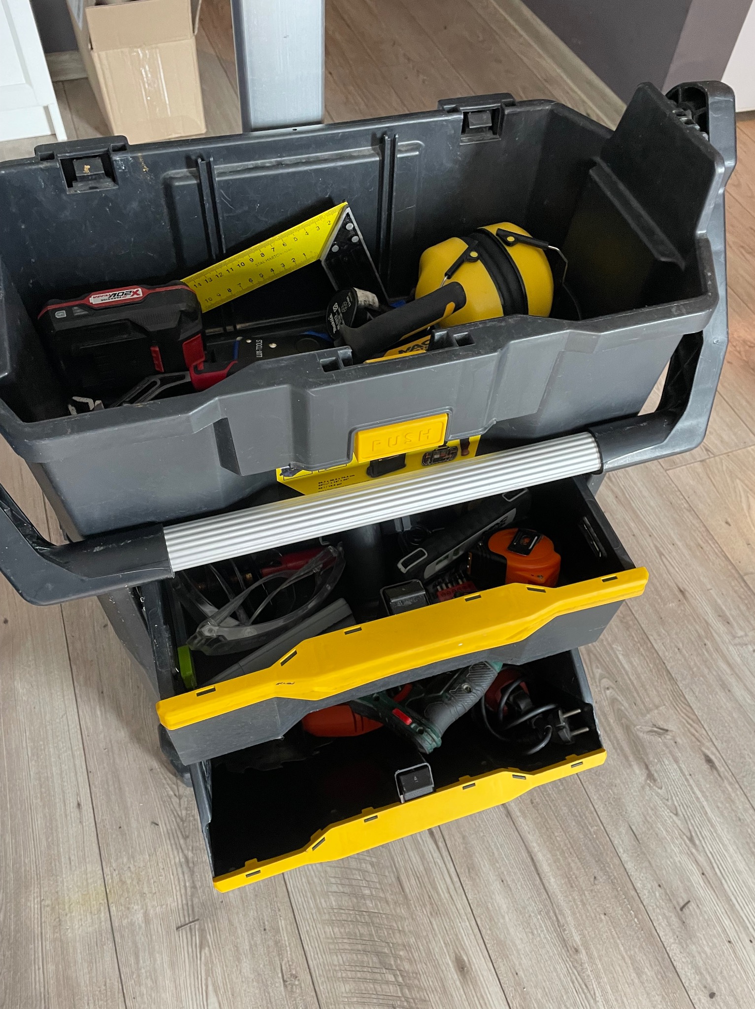 Otwarta skrzynka narzędziowa z kilkoma szufladami, wypełniona różnymi narzędziami, w tym miarką, kątownikiem, słuchawkami ochronnymi, wkrętarką akumulatorową i okularami ochronnymi.