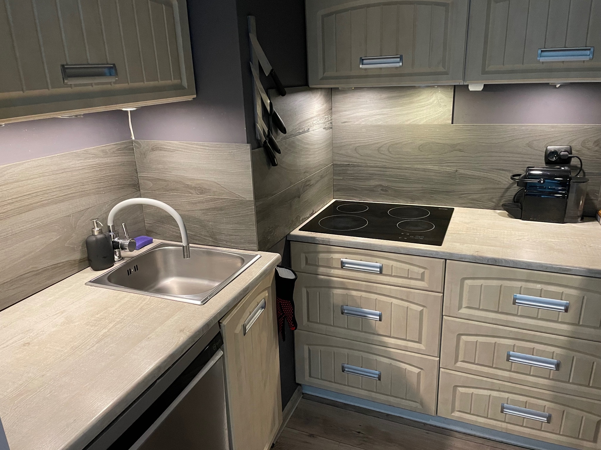 Jasna kuchnia z drewnopodobnymi frontami, widoczny zlew z baterią, płyta indukcyjna, ekspres do kawy i magnetyczny uchwyt na noże