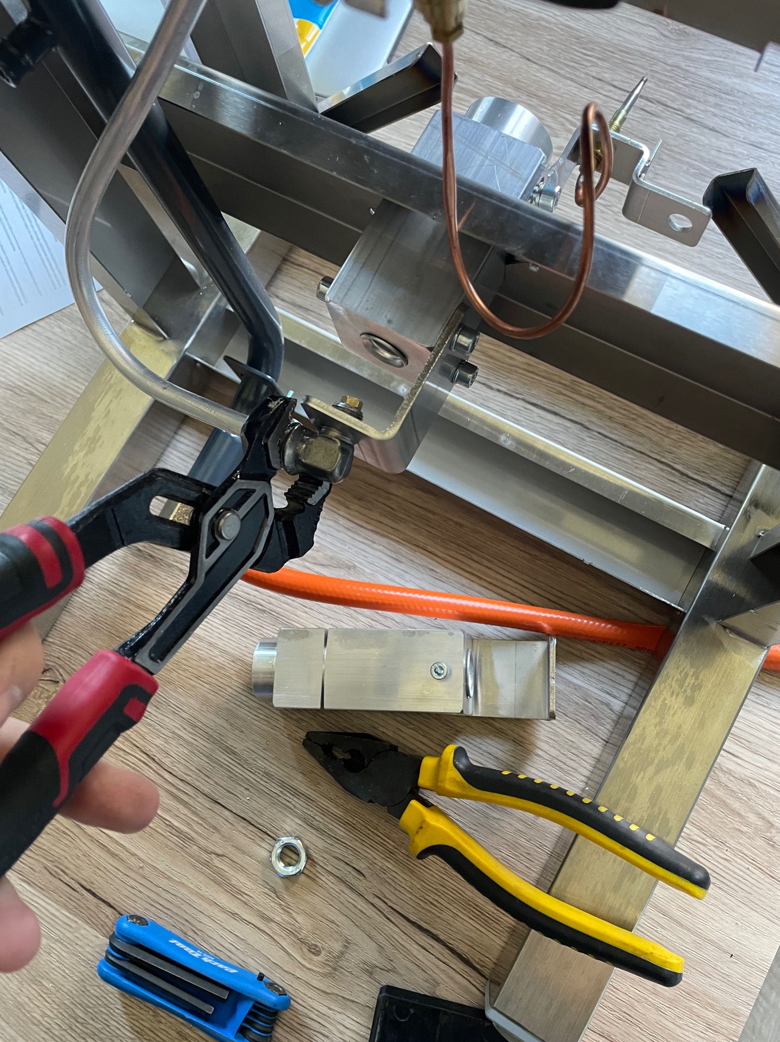 Szczypce z czerwonymi uchwytami zaciskają nakrętkę na metalowej konstrukcji, obok leżą narzędzia, w tym klucz imbusowy w niebieskiej obudowie oraz szczypce z żółtymi uchwytami.