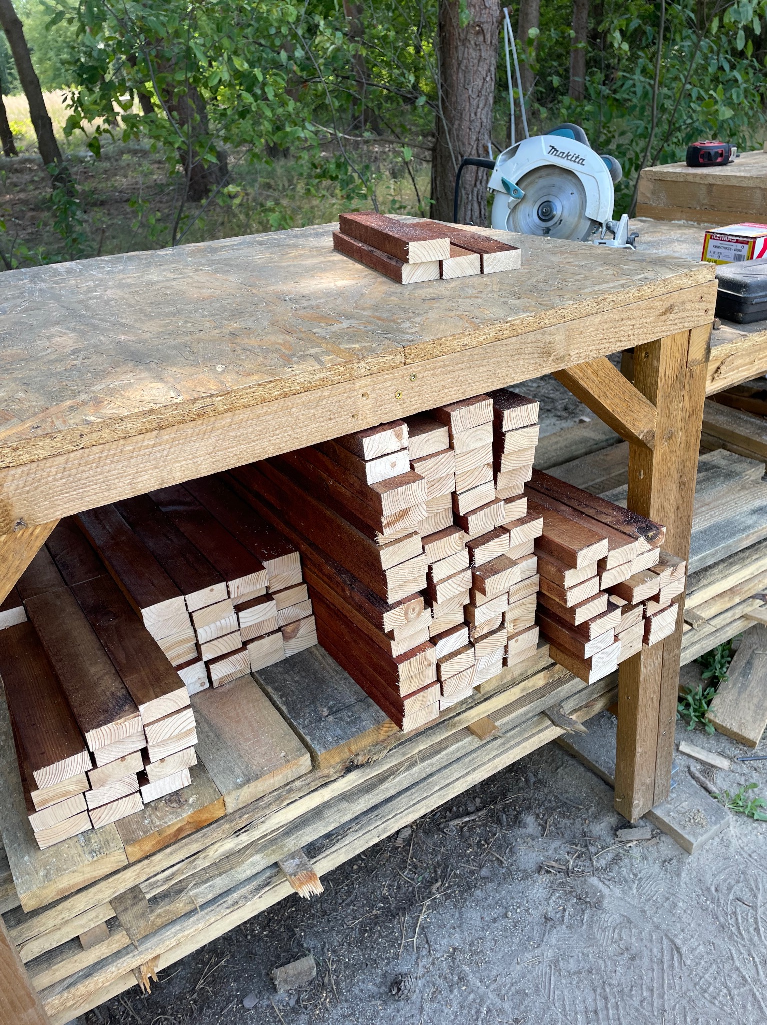 Przygotowane, przycięte i zaimpregnowane deski ułożone na drewnianym stole warsztatowym w Warszawie, z widoczną piłą tarczową Makita w tle, sugerujące etap przygotowania do prac remontowych...