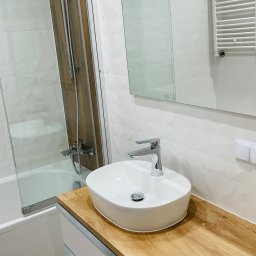 CWION SPÓŁKA Z OGRANICZONĄ ODPOWIEDZIALNOŚCIĄ - Nowoczesna łazienka z owalną umywalką nablatową na blacie z drewnopodobnym wykończeniem, białe płytki z delikatną fakturą na ścianach, przeszklona kabina prysznicowa z brązowym akcentem...