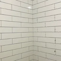 Świeżo ułożone, białe, prostokątne płytki na ścianie w rogu pomieszczenia z widocznymi krzyżykami dystansowymi, na dole kadru fragment poziomicy.