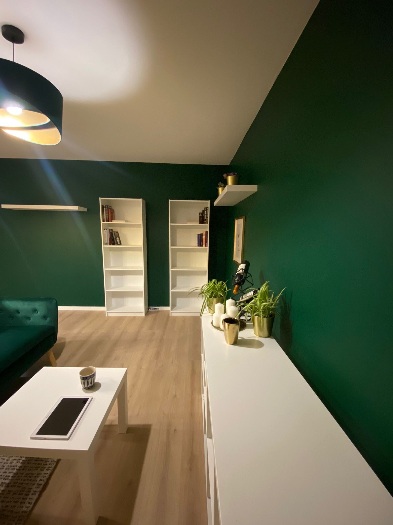 Salon z ciemnozieloną ścianą, białymi regałami i komodą, oświetlony lampą z abażurem, z elementami dekoracyjnymi jak rośliny w złotych doniczkach i tablet na białym stoliku kawowym.