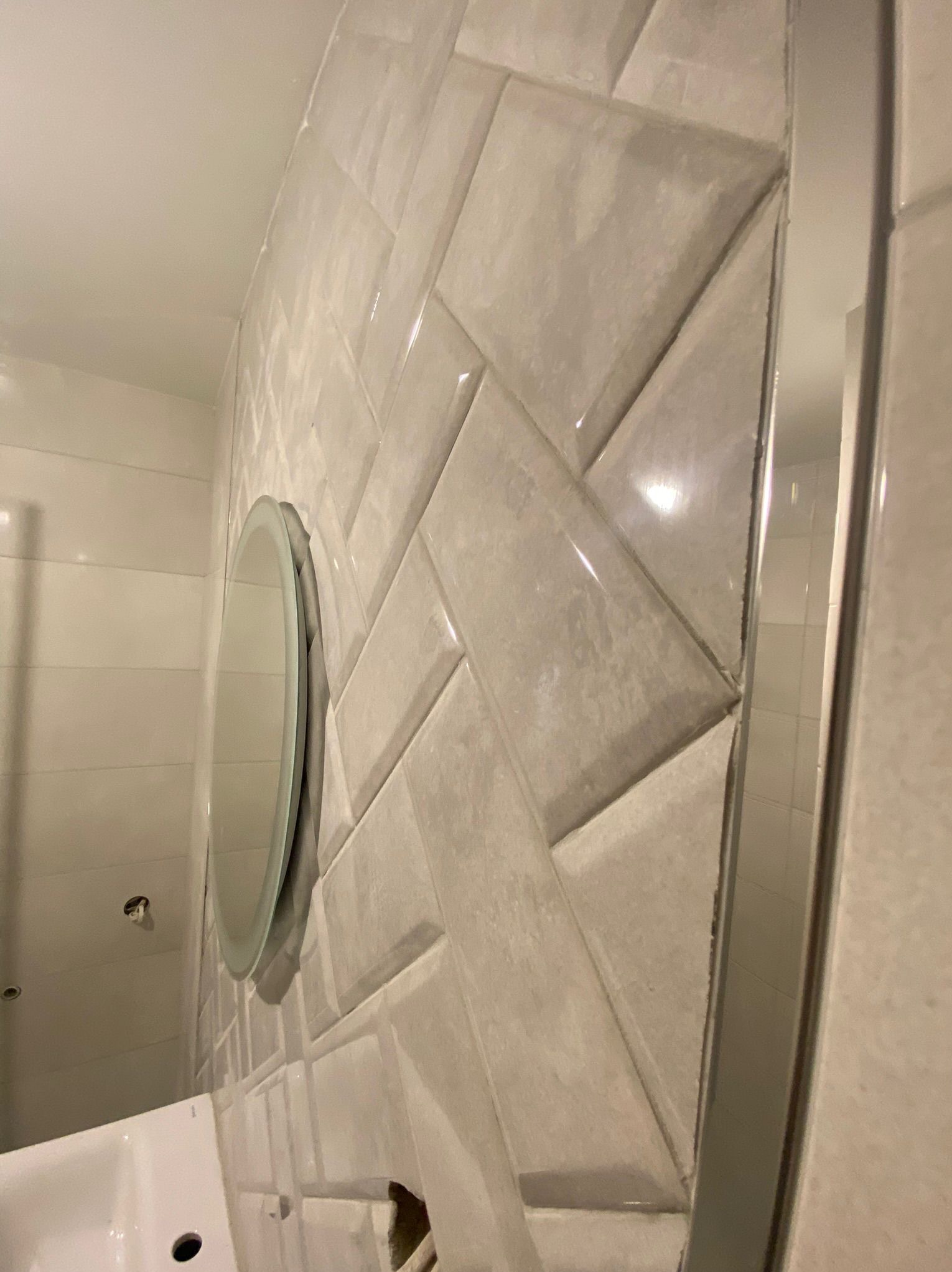 Ściana w łazience wyłożona błyszczącymi, beżowymi płytkami ułożonymi w jodełkę, okrągłe lustro z podświetleniem LED, widoczna armatura i fragment wanny.