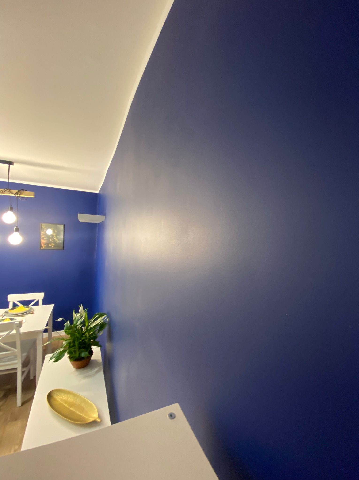 Ściana w odcieniach granatu po malowaniu, widok z perspektywy narożnika, z elementami wystroju wnętrza: biały stół z krzesłami, roślina doniczkowa i dekoracyjna misa w kolorze złotym.