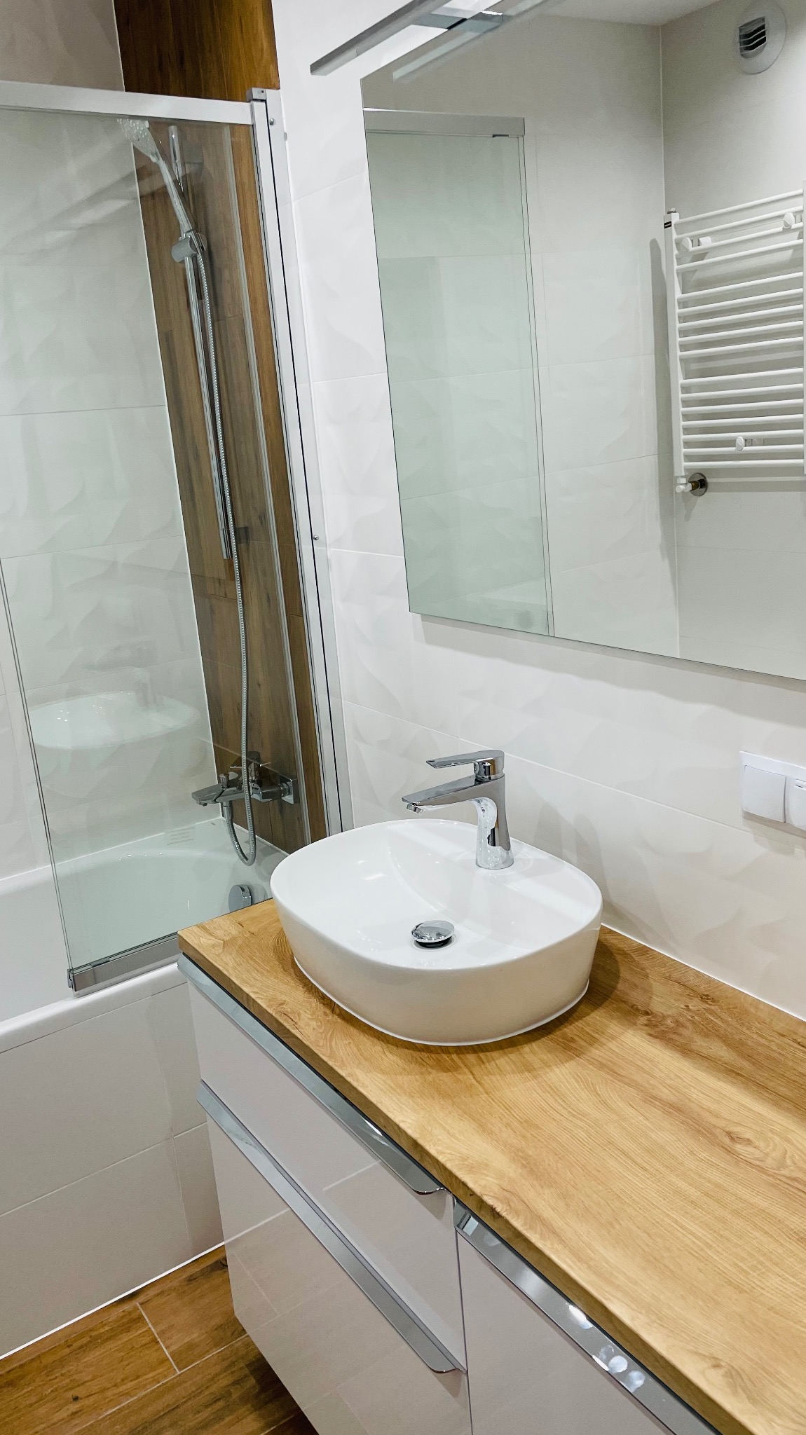 Nowoczesna łazienka z owalną umywalką nablatową na blacie z drewnopodobnym wykończeniem, białe płytki z delikatną fakturą na ścianach, przeszklona kabina prysznicowa z brązowym akcentem...