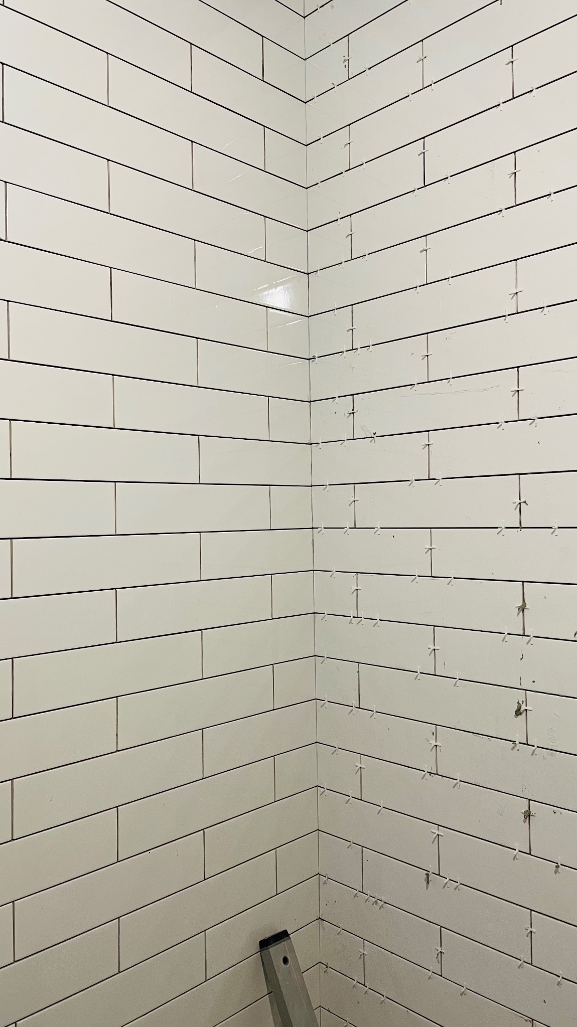 Świeżo ułożone, białe, prostokątne płytki na ścianie w rogu pomieszczenia z widocznymi krzyżykami dystansowymi, na dole kadru fragment poziomicy.