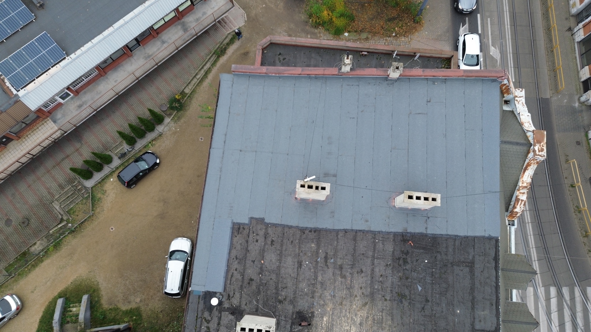 Widok z góry na płaski dach budynku z widocznymi kominami wentylacyjnymi, częściowo pokryty nową szarą papą, a częściowo starą, czarną papą z uszkodzeniami, z fragmentem rynny wzdłuż krawędzi dachu...