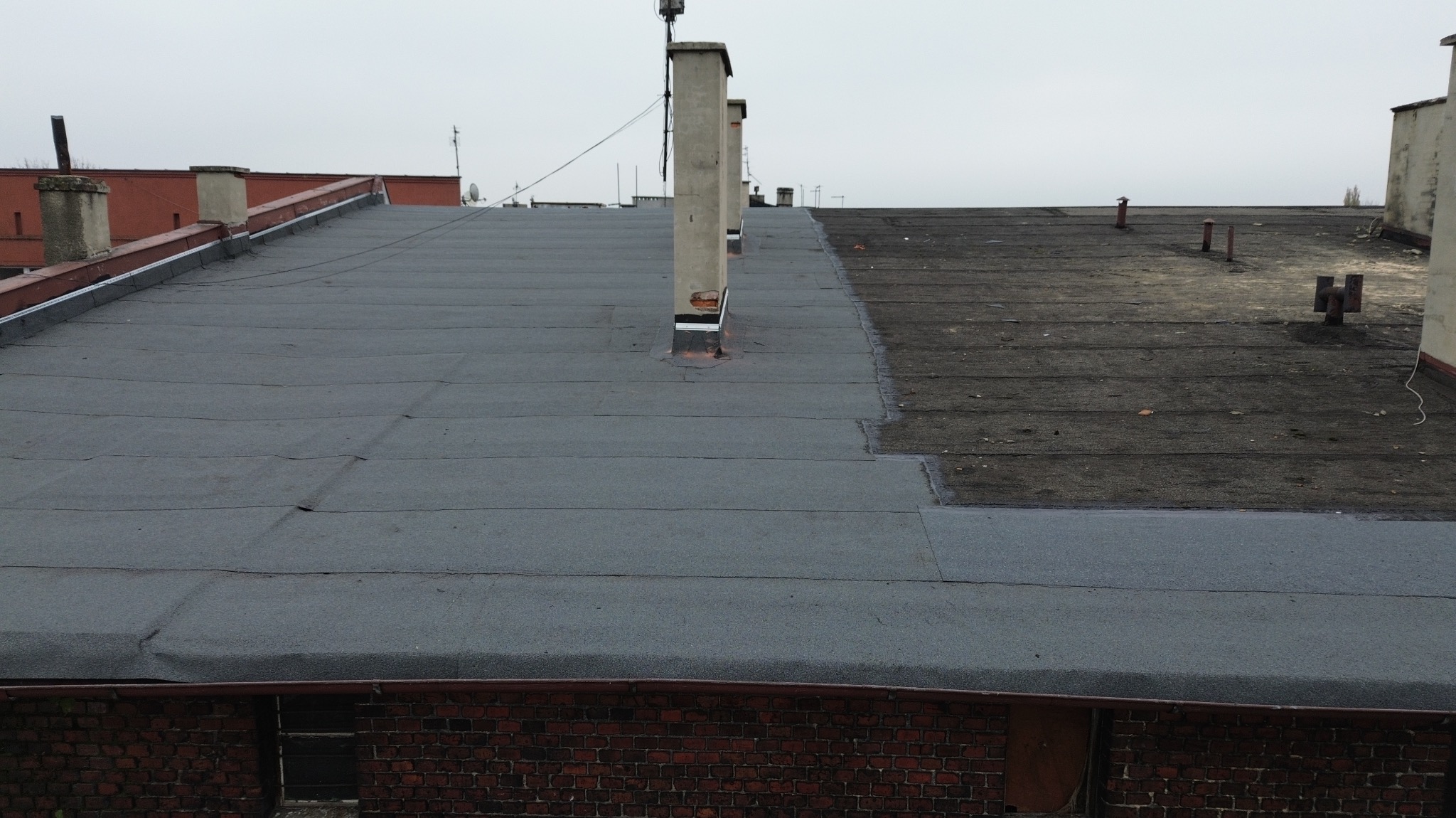 Płaski dach budynku pokryty częściowo nową, szarą papą termozgrzewalną, a częściowo starą, ciemną papą. Widoczne kominy i elementy wentylacyjne. Fragment muru z czerwonej cegły w dolnej części kadru.