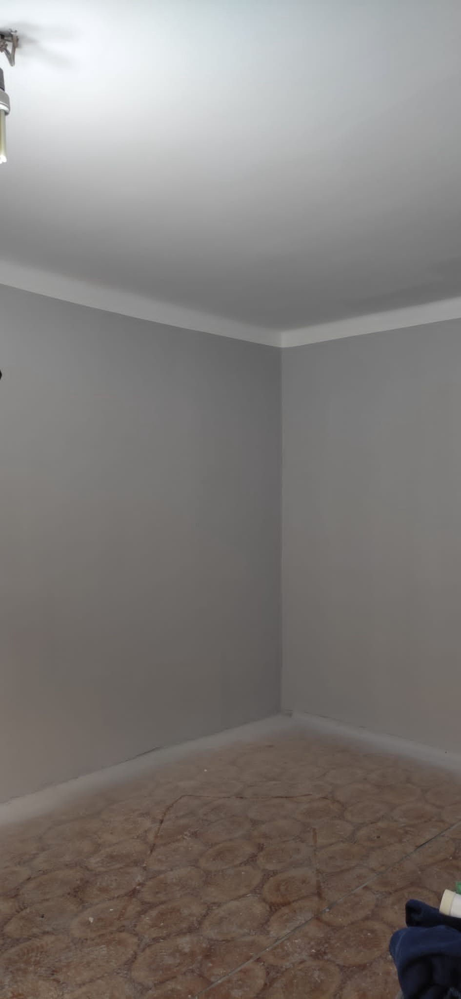 Świeżo pomalowane szare ściany z białym wykończeniem sufitu w rogu pokoju, widoczna część podłogi z brązową mozaiką.