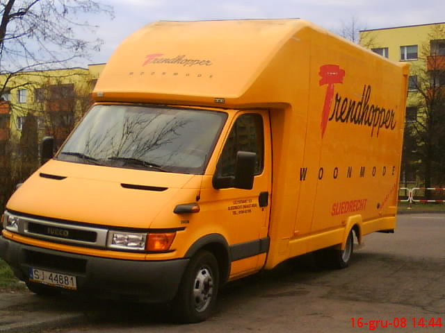 Żółty samochód dostawczy marki Iveco z napisem 'Trendhopper Woonmode Sliedrecht' na tle bloku mieszkalnego.