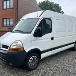 Biały Renault Master, zaparkowany na żwirowym podłożu przed ceglanym budynkiem z ciemnymi oknami, z metalowym ogrodzeniem w tle.