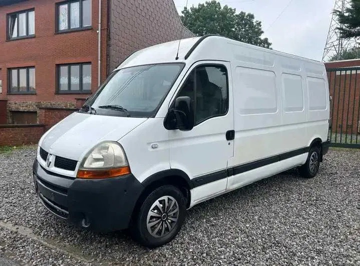 Biały Renault Master, zaparkowany na żwirowym podłożu przed ceglanym budynkiem z ciemnymi oknami, z metalowym ogrodzeniem w tle.