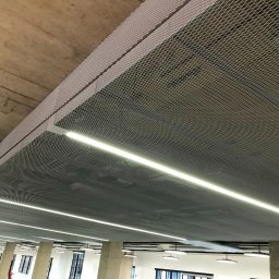 BUILDERS GROUP SPÓŁKA Z OGRANICZONĄ ODPOWIEDZIALNOŚCIĄ - Metalowa siatka sufitowa z wbudowanym oświetleniem LED w przestrzeni biurowej, widoczne betonowe elementy konstrukcyjne i kolumny.