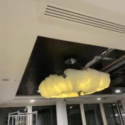 Niedokończone prace sufitowe z widocznymi elementami instalacji elektrycznej i dekoracyjną lampą w kształcie chmury, drabina w tle, fragment wentylacji sufitowej z widocznymi śladami szpachlowania.