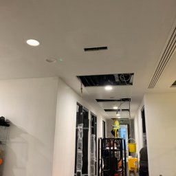 BUILDERS GROUP SPÓŁKA Z OGRANICZONĄ ODPOWIEDZIALNOŚCIĄ - Korytarz biurowy w trakcie prac remontowych sufitu podwieszanego, widoczny pracownik na podnośniku koszowym, otwarte przestrzenie w suficie ukazujące instalacje, zarysowane miejsca pod nowe...