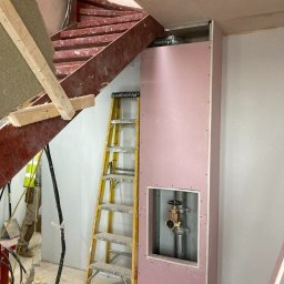 BUILDERS GROUP SPÓŁKA Z OGRANICZONĄ ODPOWIEDZIALNOŚCIĄ - Montaż obudowy z różowej płyty gipsowej wokół rur z zaworem, widoczna żółta drabina i fragment schodów spiralnych w surowym stanie.