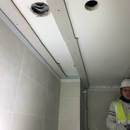 BUILDERS GROUP SPÓŁKA Z OGRANICZONĄ ODPOWIEDZIALNOŚCIĄ - Montaż płyty karton gips na suficie w łazience z wyciętymi otworami na oświetlenie, widoczny glazurnik w kasku i kamizelce odblaskowej.