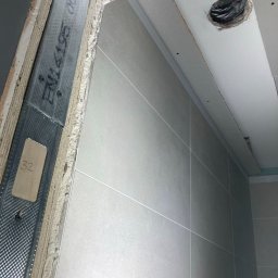 BUILDERS GROUP SPÓŁKA Z OGRANICZONĄ ODPOWIEDZIALNOŚCIĄ - Wnętrze w trakcie wykańczania: widok na szkielet metalowy przy otworze drzwiowym, fragment ściany wyłożonej szarymi płytkami oraz otwór w suficie z zabezpieczonymi kablami.