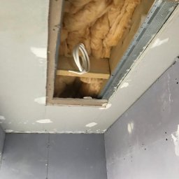 BUILDERS GROUP SPÓŁKA Z OGRANICZONĄ ODPOWIEDZIALNOŚCIĄ - Otwór w suficie z widoczną izolacją z wełny mineralnej i przewodami elektrycznymi, ściany wykończone płytami kartonowo-gipsowymi przed szpachlowaniem.