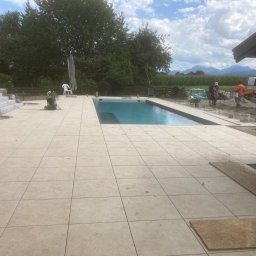 BUILDERS GROUP SPÓŁKA Z OGRANICZONĄ ODPOWIEDZIALNOŚCIĄ - Układanie jasnych, kwadratowych płytek wokół basenu na zewnątrz; w tle widoczni pracownicy i krajobraz z górami.