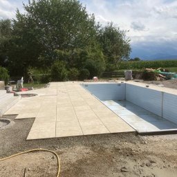 BUILDERS GROUP SPÓŁKA Z OGRANICZONĄ ODPOWIEDZIALNOŚCIĄ - Etap budowy basenu: układanie beżowych płyt wokół niecki, widoczny pracownik w okularach ochronnych, w tle koparka i sterta materiałów budowlanych.