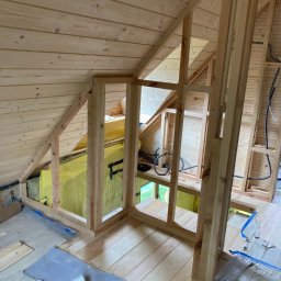 BUILDERS GROUP SPÓŁKA Z OGRANICZONĄ ODPOWIEDZIALNOŚCIĄ - Drewniana konstrukcja ścianki działowej w trakcie ocieplania poddasza, widoczna żółta wełna mineralna, częściowo zabezpieczona folią budowlaną.