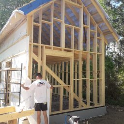 BUILDERS GROUP SPÓŁKA Z OGRANICZONĄ ODPOWIEDZIALNOŚCIĄ - Wznoszenie szkieletowej konstrukcji drewnianej domu z widocznym ociepleniem ścian zewnętrznych, pracownik w koszulce z logo firmy budowlanej oraz kompresor powietrza na placu budowy.