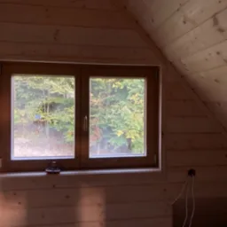Drewniane wykończenie poddasza z oknem w miejscowości Dobra, widok na jesienny las, widoczne kable wystające ze ściany i pudełko z narzędziami na parapecie.