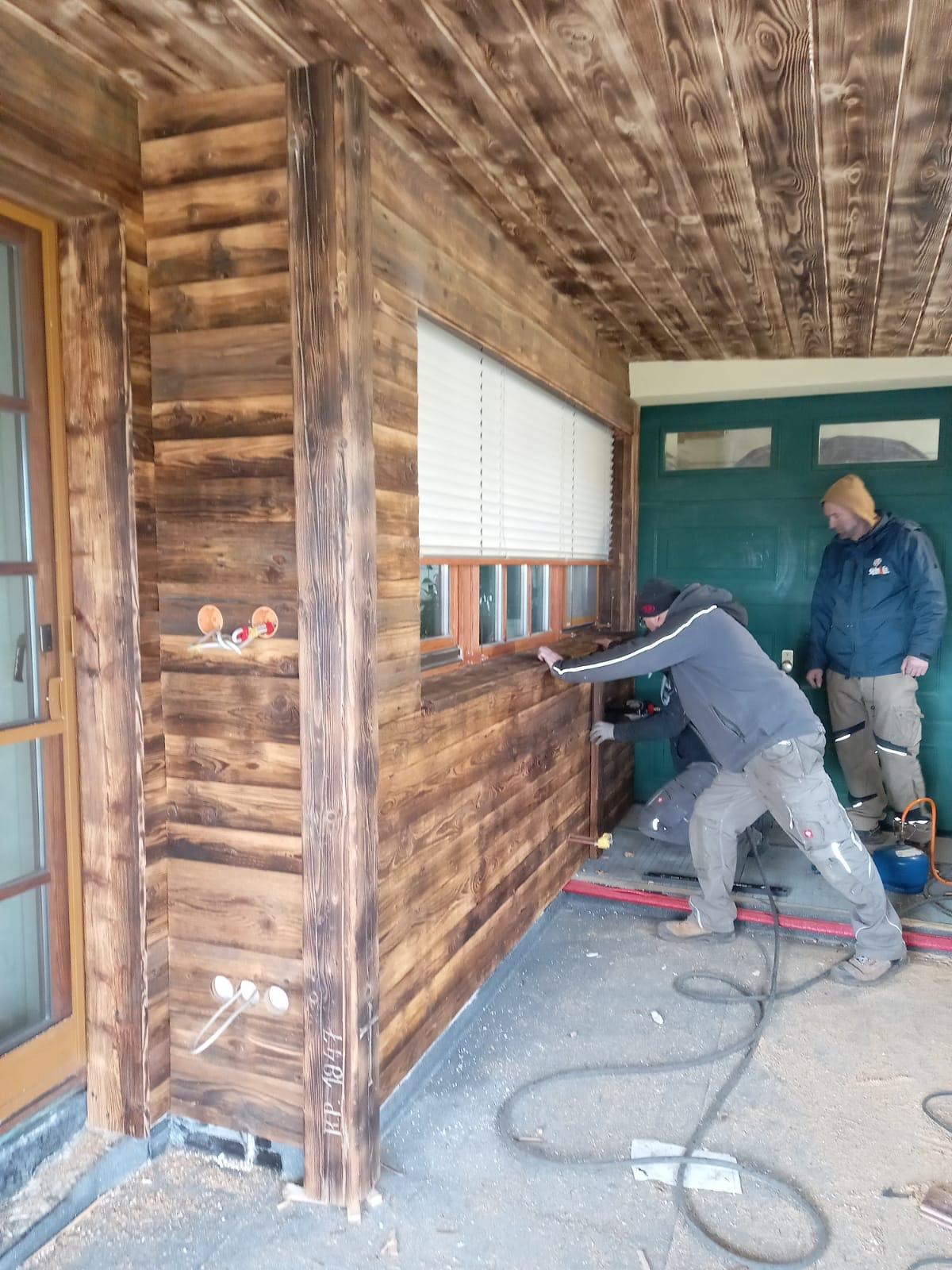 Monteraż drewnianej boazerii na ścianie zewnętrznej budynku; widoczne narzędzia, przewody oraz dwóch pracowników w trakcie pracy. Na ścianie widoczne otwory na instalacje.