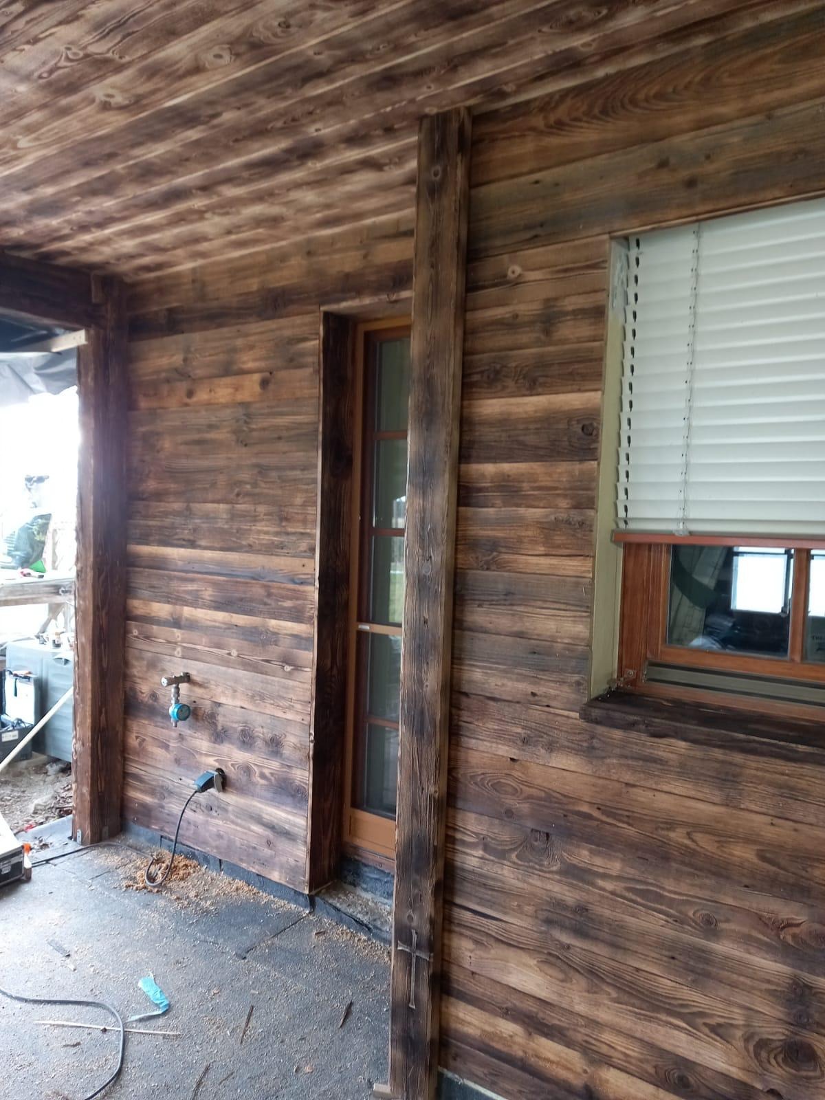 Drewniana elewacja domu z oknem z żaluzjami, widoczny fragment podbitki dachowej, instalacja wodna z podłączonym kablem elektrycznym oraz narzędzia na podłodze.