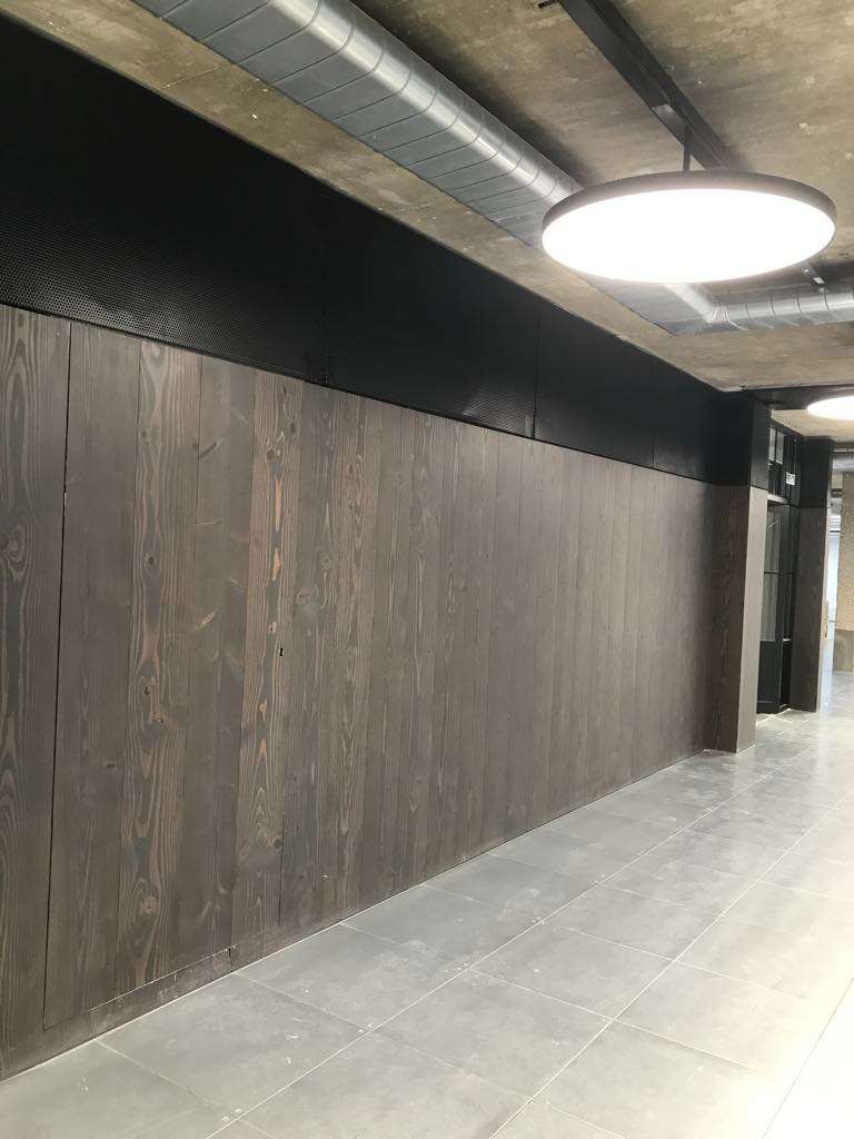 Wykończona ściana z ciemnego drewna z widocznym rysunkiem słojów, zamontowana w biurze w miejscowości Dobra, z szarą podłogą i widocznymi elementami instalacji pod sufitem.