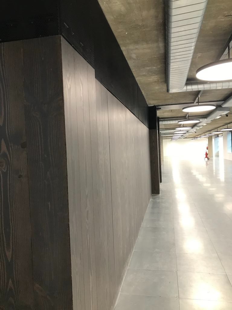 Długa ściana wykończona panelami z ciemnego drewna, przechodząca w jaśniejsze panele, z widocznymi elementami instalacji pod sufitem i błyszczącą podłogą w przestronnym biurze.