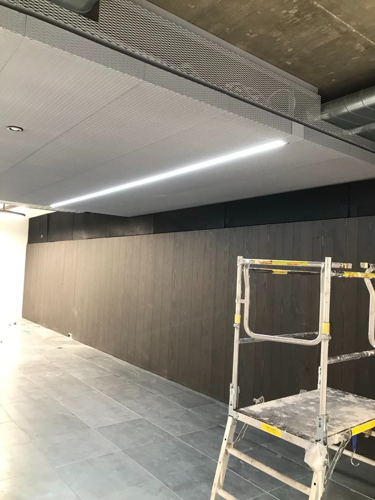 Wnętrze biurowe w trakcie wykańczania: ciemne panele ścienne z widoczną strukturą drewna, metalowa siatka sufitowa z oświetleniem liniowym LED, szare płytki podłogowe, rusztowanie malarskie...