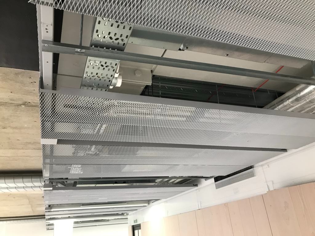 Sufit podwieszany z metalowej siatki o wzorze romboidalnym, widoczne elementy instalacji wentylacyjnej i oświetleniowej, surowy betonowy strop w tle.