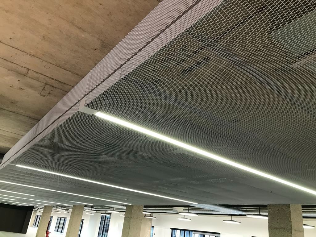 Metalowa siatka sufitowa z wbudowanym oświetleniem LED w przestrzeni biurowej, widoczne betonowe elementy konstrukcyjne i kolumny.