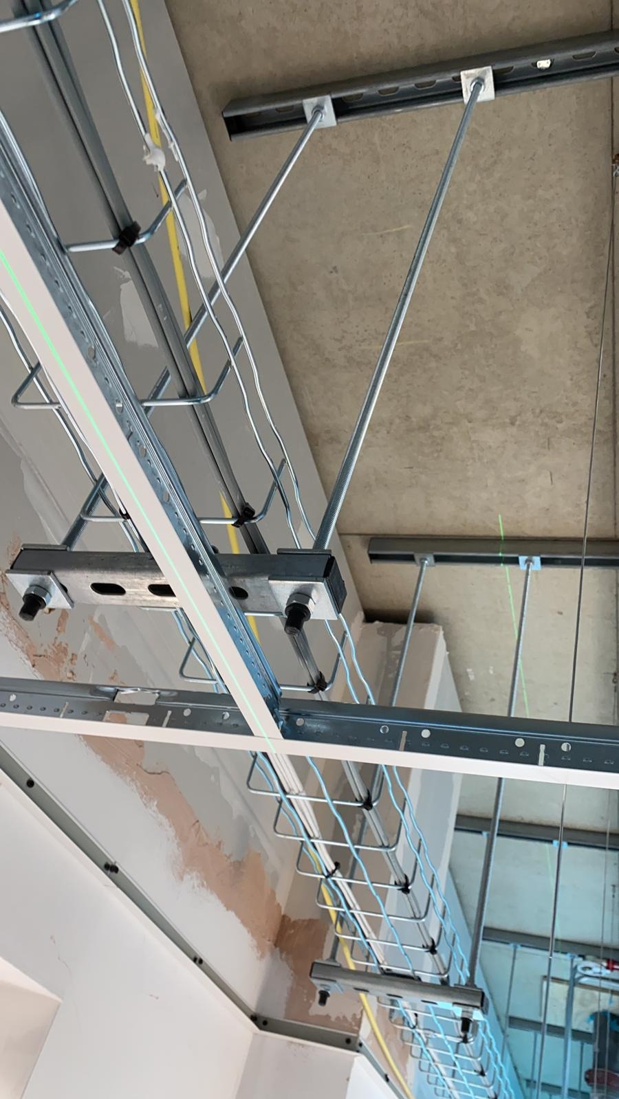 Metalowy stelaż sufitowy z profilami i przewodami elektrycznymi, widoczne elementy mocujące i laserowy poziom wyznaczający linię, surowy betonowy strop w tle.