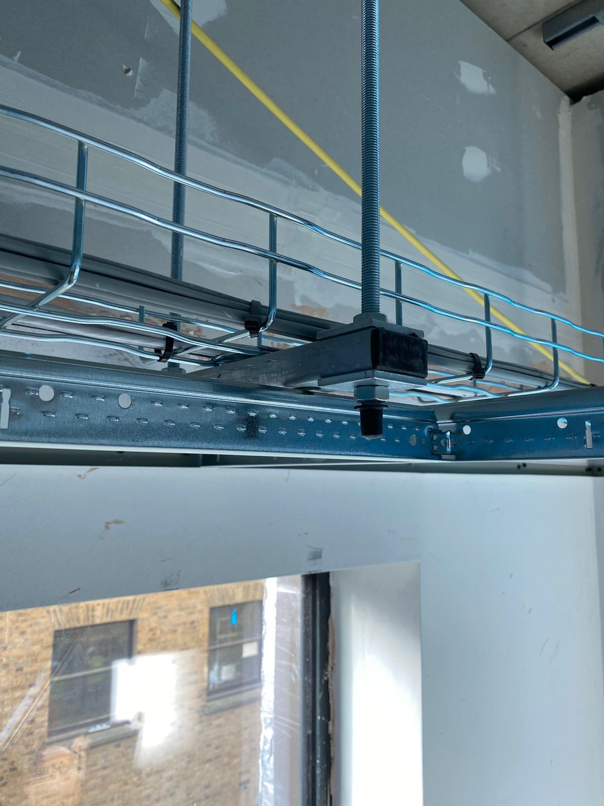 Metalowa konstrukcja podwieszona do sufitu z widocznymi przewodami elektrycznymi, mocowana na gwintowanych prętach w pomieszczeniu z oknem i ścianami przygotowanymi do wykończenia.